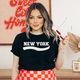 A woman wearing a black t-shirt with 'NEW YORK' printed in white across the chest, paired with a red and white checkered skirt, holding red-framed sunglasses in a brightly lit indoor setting.