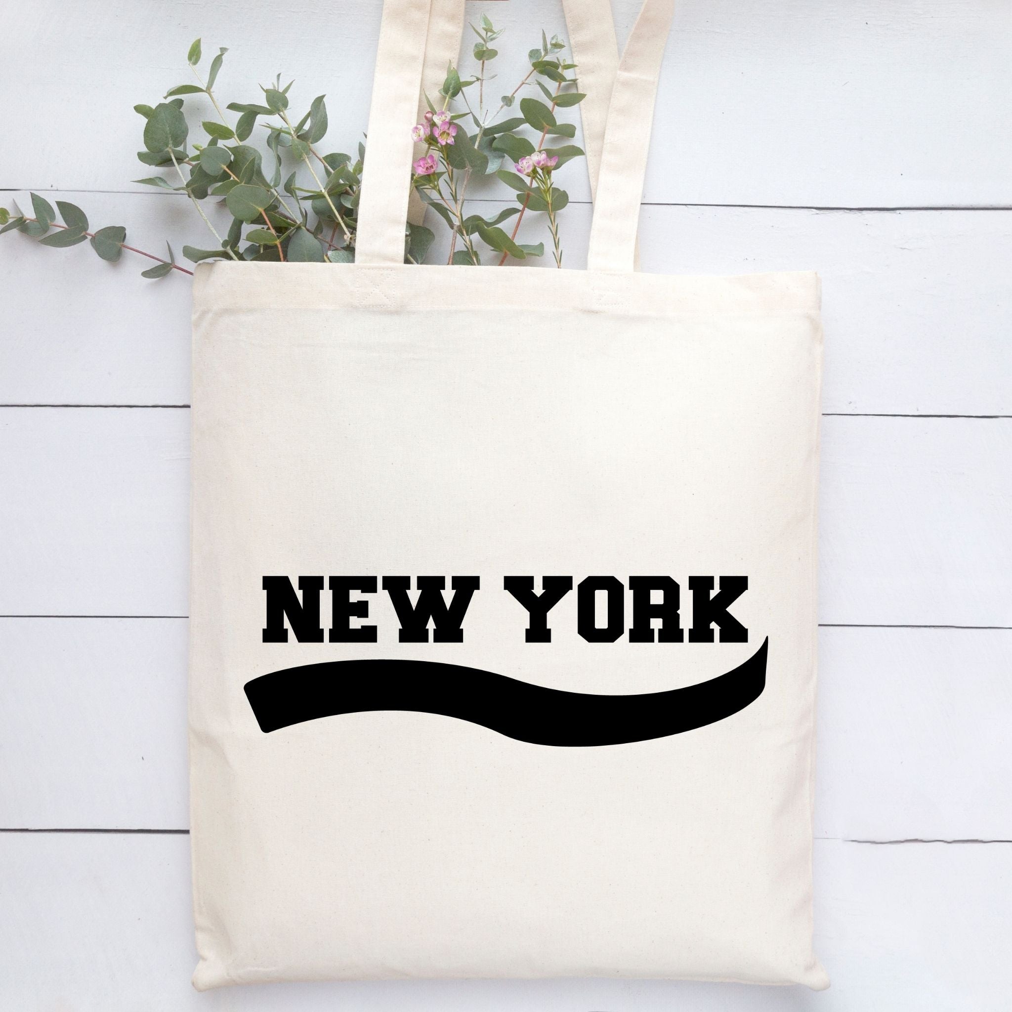 An off-white canvas tote bag with 'NEW YORK' printed in bold black letters and a black wavy line underneath, styled on a white wood plank background with green eucalyptus stems.