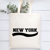 An off-white canvas tote bag with 'NEW YORK' printed in bold black letters and a black wavy line underneath, styled on a white wood plank background with green eucalyptus stems.