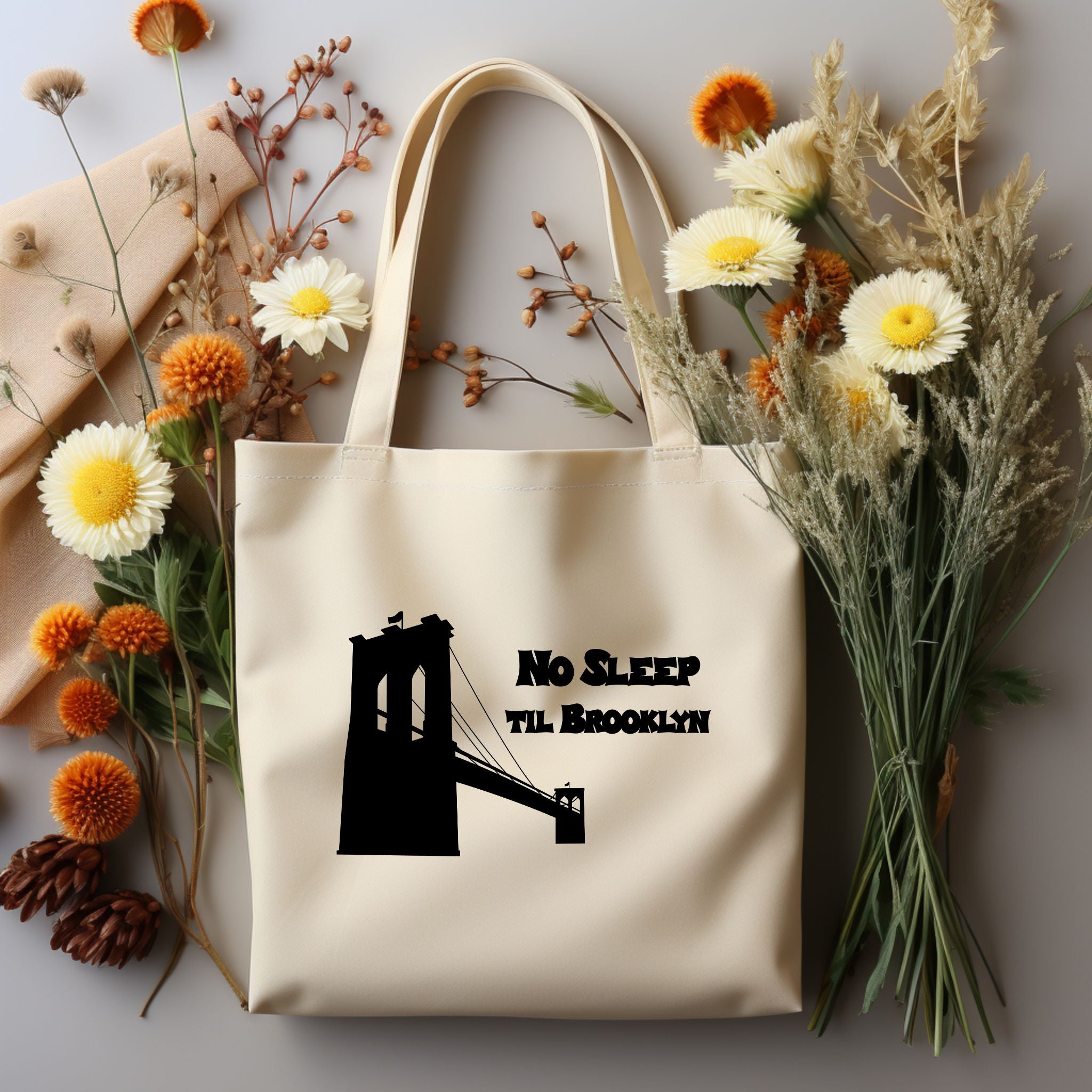 A cream canvas tote bag with a black graphic of the Brooklyn Bridge and the text 'No Sleep Til Brooklyn,' styled in a flat lay with dried flowers, pinecones, and a beige scarf on a light gray background.