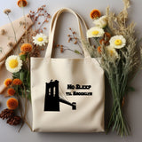 A cream canvas tote bag with a black graphic of the Brooklyn Bridge and the text 'No Sleep Til Brooklyn,' styled in a flat lay with dried flowers, pinecones, and a beige scarf on a light gray background.
