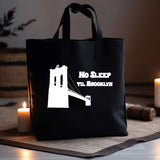 A black canvas tote bag with a white graphic of the Brooklyn Bridge and the text 'NO SLEEP TIL BROOKLYN' is displayed on a woven mat with candles in a cozy indoor setting.
