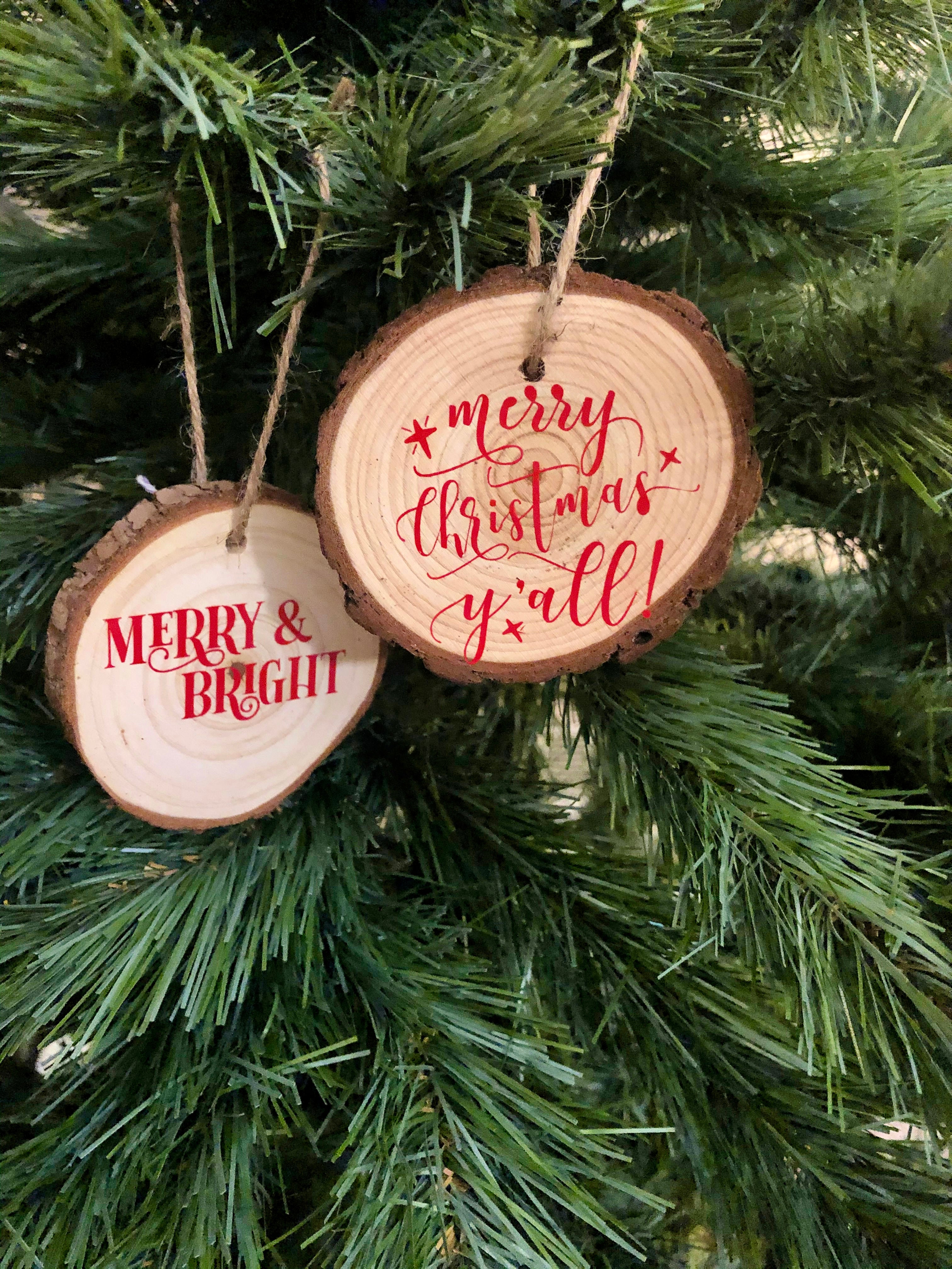 Two round wooden Christmas tree ornaments hanging on a green artificial tree. One ornament is a natural wood slice with the words 'MERRY & BRIGHT' in red, and the other is a darker wood slice with 'merry christmas y'all!' in red script.