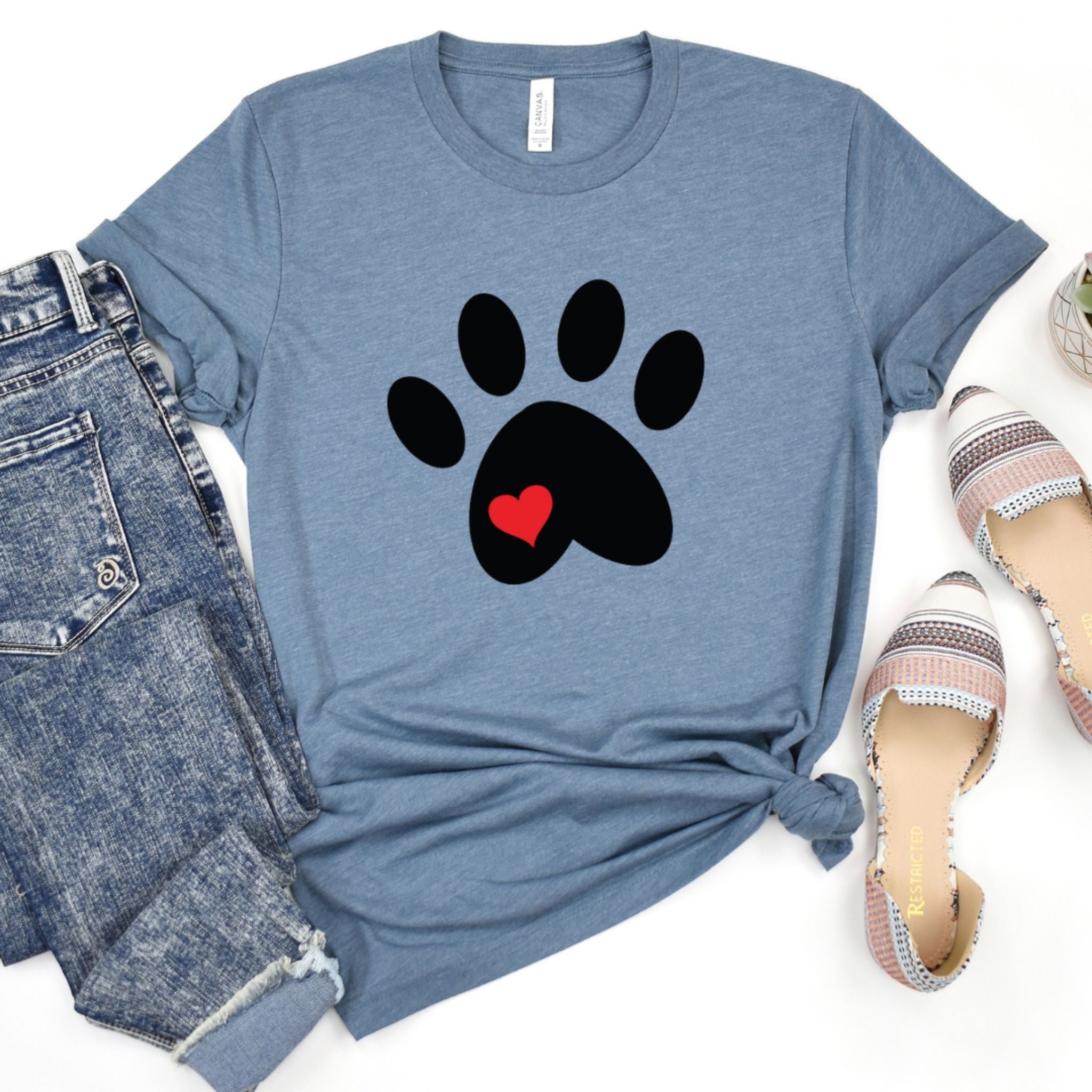 A heathered blue short-sleeve T-shirt laid flat with a black paw print graphic and a small red heart in the center. The shirt is styled in a flat lay with blue jeans on the left and patterned slip-on shoes on the right against a white background.
