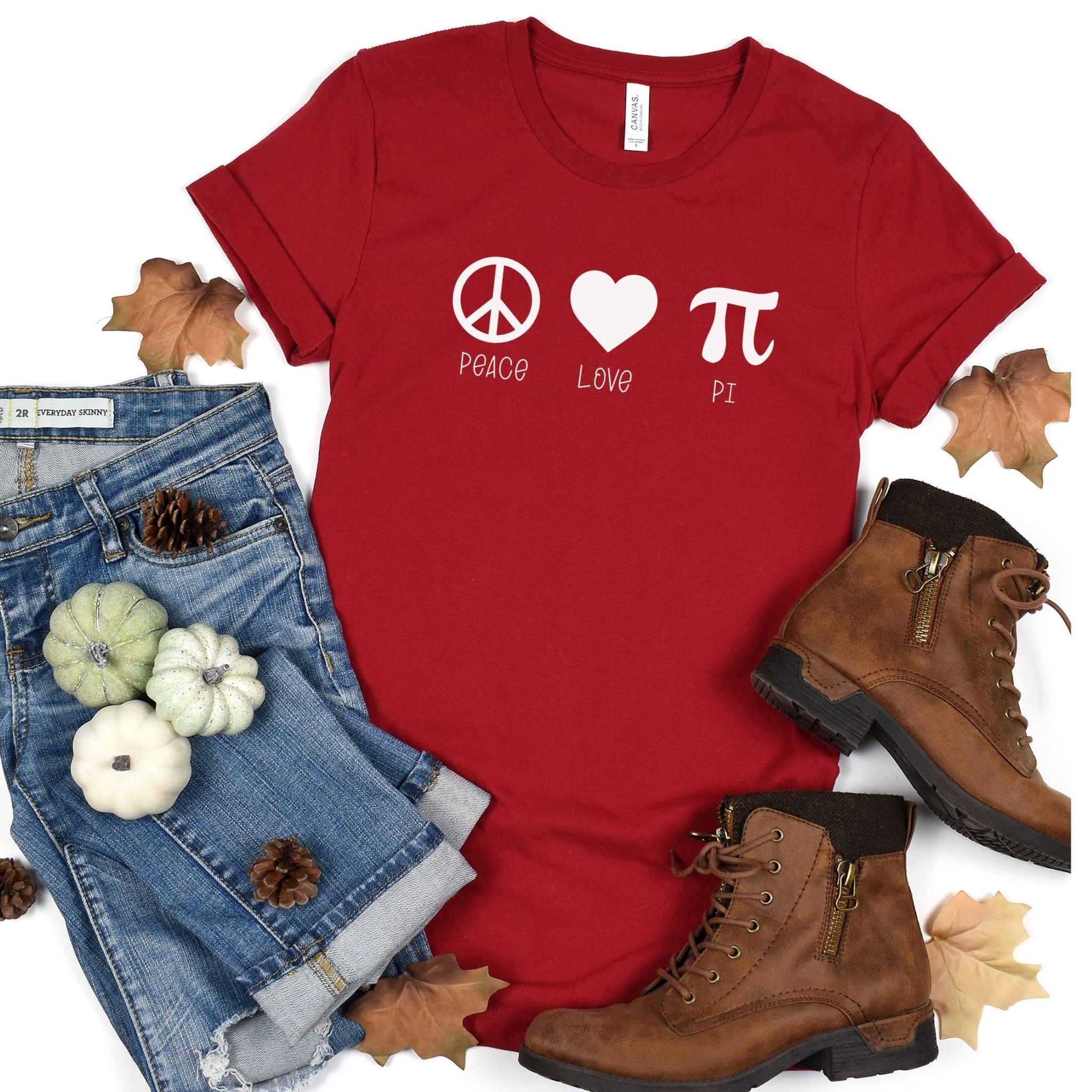 Red shirt with white Peace Love Pi wititen.  Jeans to the left with pumpkins in white and green, fall leaves, and brown boots to the right.