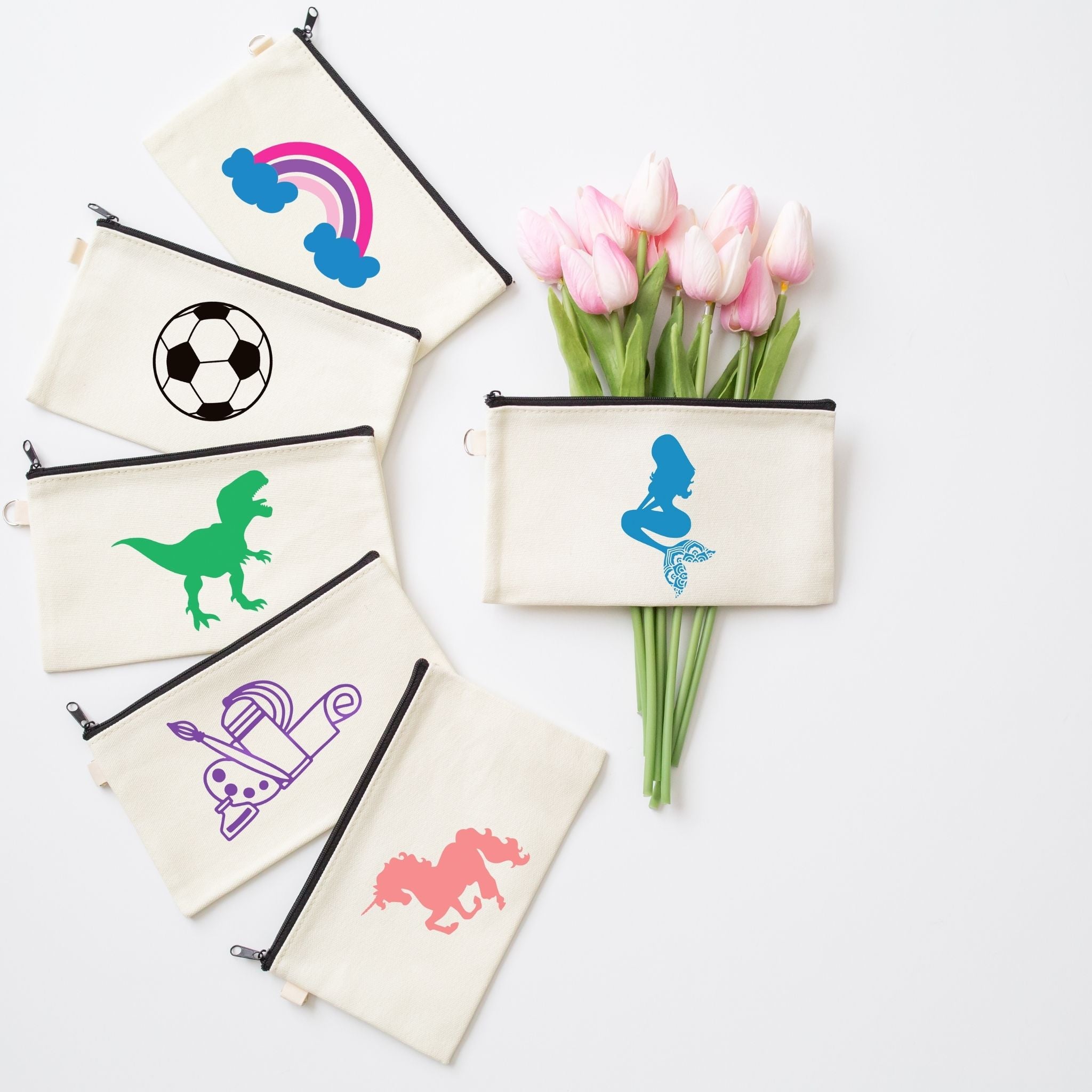 A set of cream canvas zippered pouches with black zippers, each printed with a different colorful graphic (rainbow, soccer ball, dinosaur, roller skate, bicycle, horse). A matching pouch with a blue mermaid graphic is shown next to a bouquet of pink tulips on a white background.