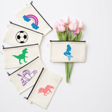 A set of cream canvas zippered pouches with black zippers, each printed with a different colorful graphic (rainbow, soccer ball, dinosaur, roller skate, bicycle, horse). A matching pouch with a blue mermaid graphic is shown next to a bouquet of pink tulips on a white background.