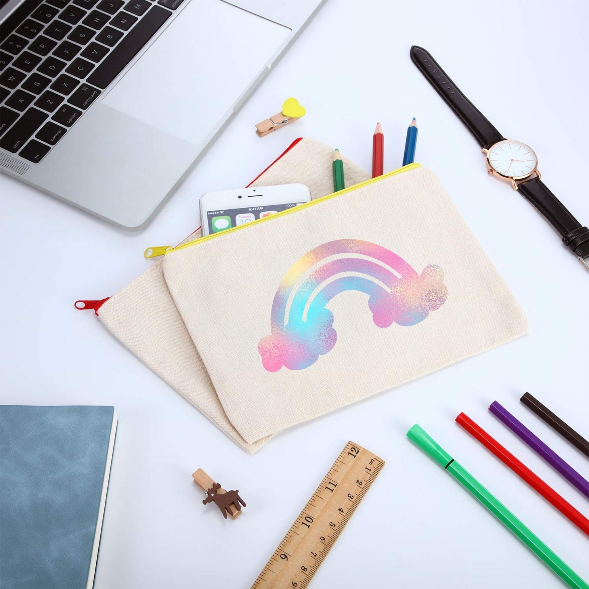 A beige canvas pencil pouch with a pastel rainbow and cloud print, styled on a white desk with a laptop corner, watch, colored pencils, a ruler, and other stationery.