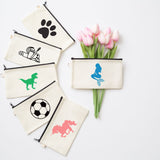 Five cream canvas zip pouches with black zippers are arranged on a white background. One pouch is closed and shows a blue mermaid graphic, while the other four are open, displaying black, green, white, and pink graphics.