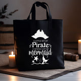 A black canvas tote bag with white printed text reading 'A Little Bit of Pirate, A Whole lot of mermaid' and small mermaid illustrations, displayed on a table with candles in a cozy indoor setting.