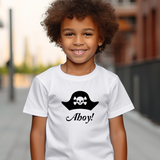 A young child with curly brown hair wearing a white t-shirt with a black pirate hat and skull graphic above the text 'Aho!'. The background is a blurred outdoor street scene.