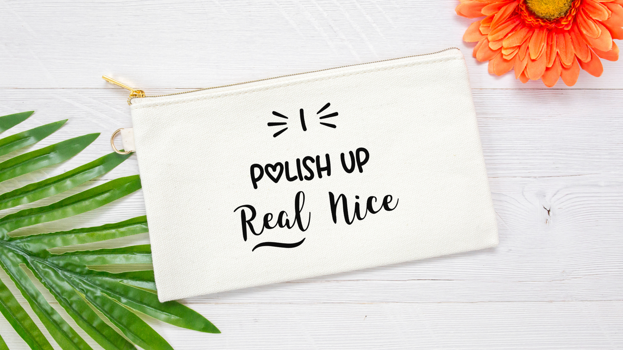 A cream canvas cosmetic pouch with black text reading 'POLISH UP Real Nice' and a small gold zipper, styled on a white wood background with green leaves and an orange flower.