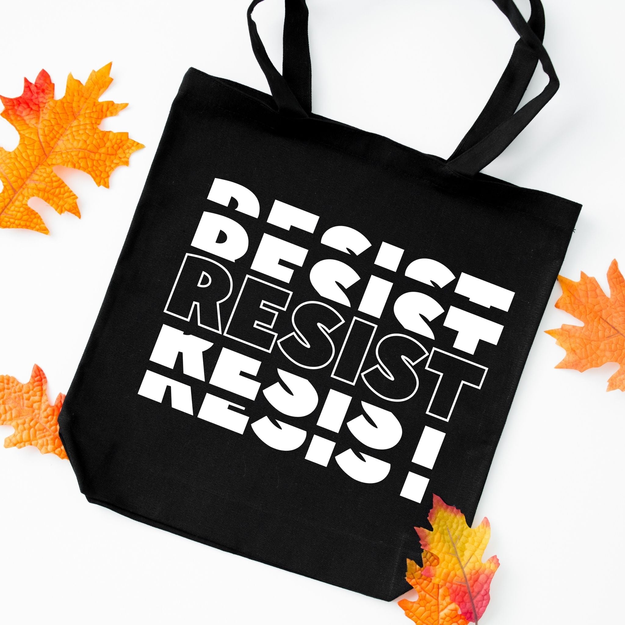 A black canvas tote bag with bold white text reading 'DESIST RESIST' repeated diagonally, styled on a white background with scattered orange and yellow autumn leaves around it.