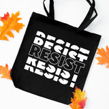 A black canvas tote bag with bold white text reading 'DESIST RESIST' repeated diagonally, styled on a white background with scattered orange and yellow autumn leaves around it.