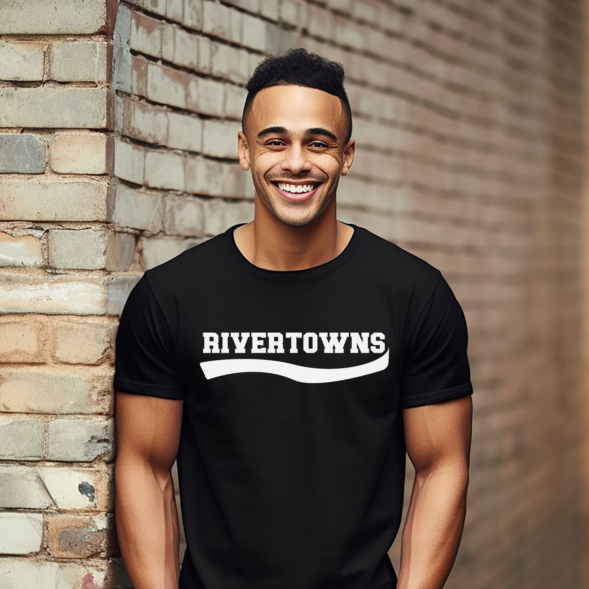 A smiling man wearing a black short-sleeve t-shirt with 'RIVERTOWNS' printed in white across the chest, standing against a brick wall.
