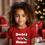 A young girl with curly brown hair smiles while wearing a red t-shirt that says 'Santa's little Helper' in white text with a small green Santa hat graphic. The background shows a cozy Christmas setting with wrapped gifts and a blurred tree with warm lights.