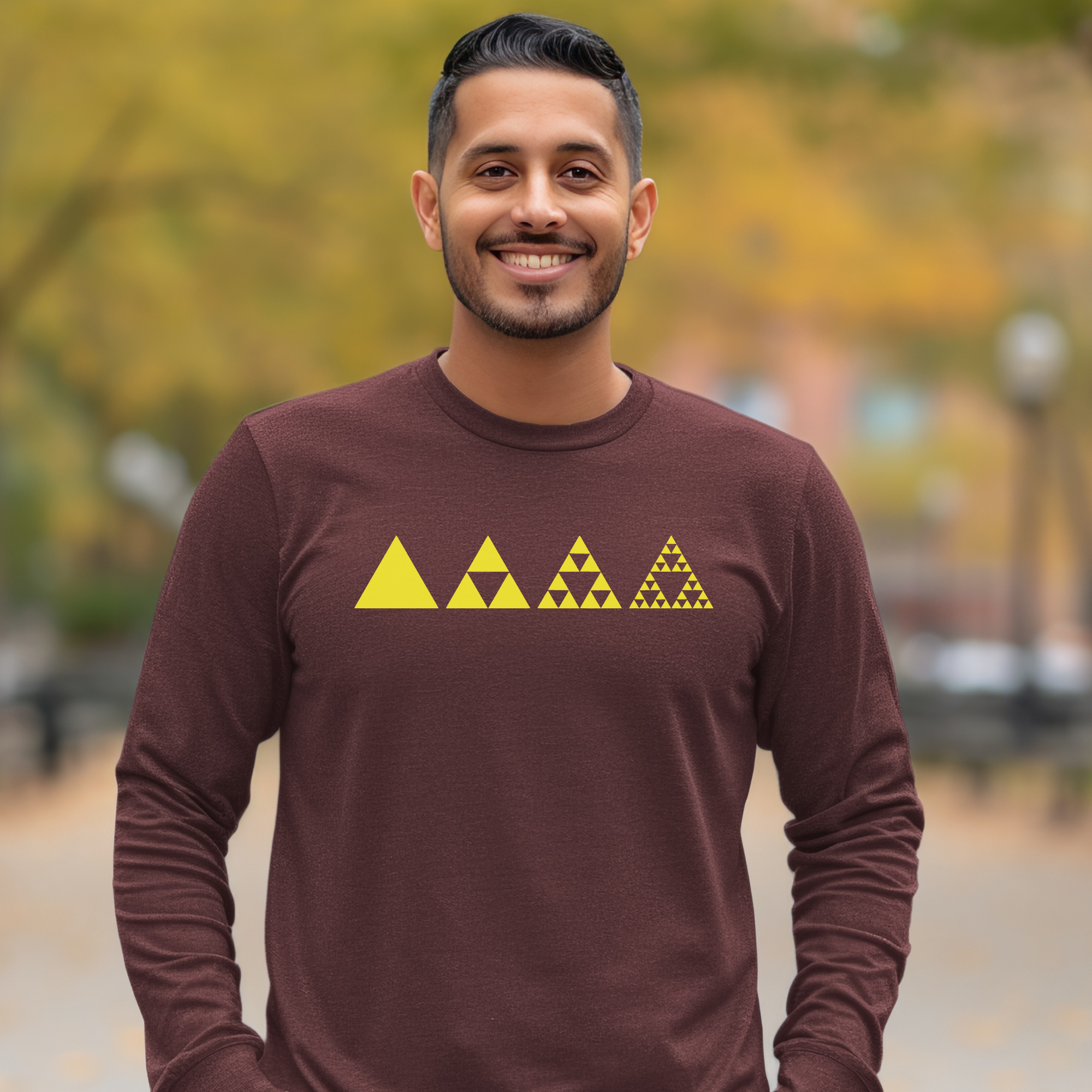 A smiling man wearing a long-sleeve maroon shirt with a yellow fractal triangle graphic across the chest, set against a blurred outdoor background of autumn trees.