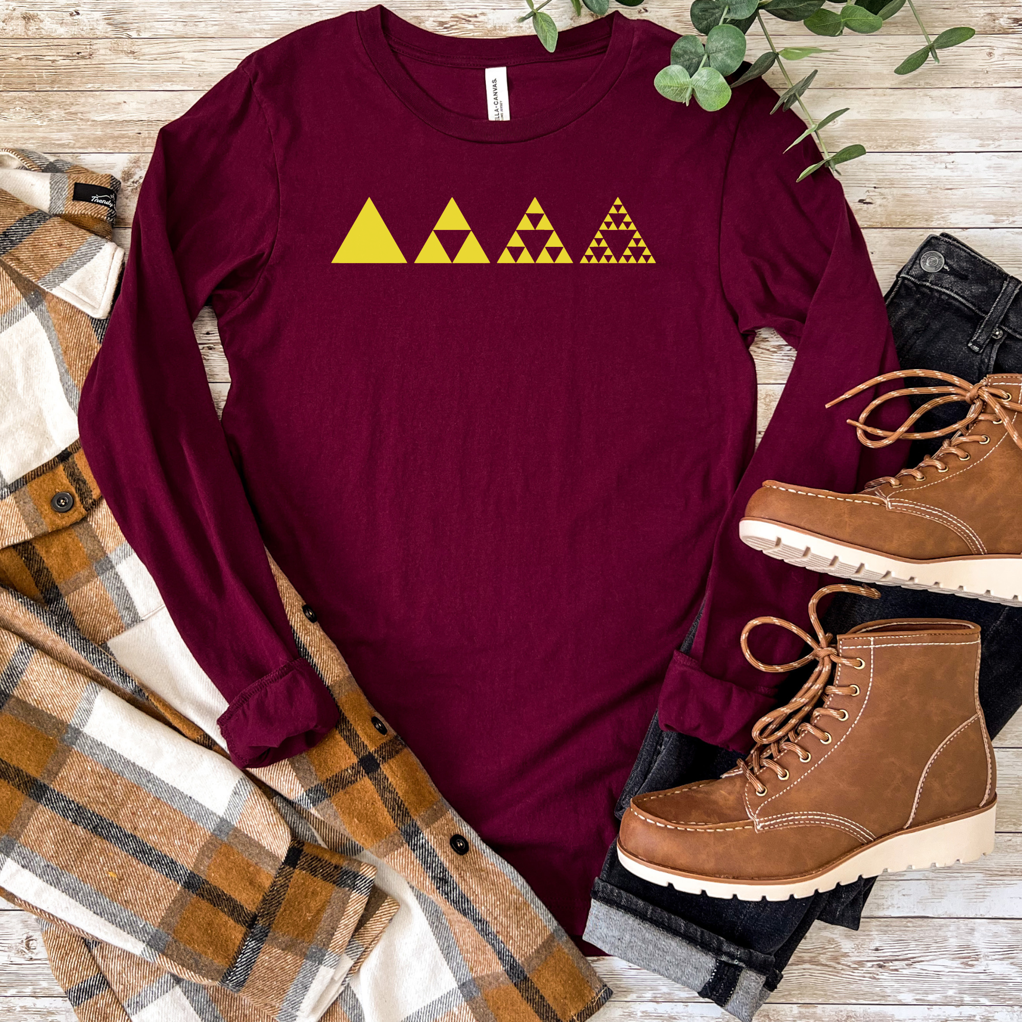 A long-sleeve maroon shirt laid flat on a rustic wood surface, featuring a yellow triangle pattern across the chest. The shirt is styled with a plaid flannel shirt, dark jeans, brown lace-up boots, and green leaves.