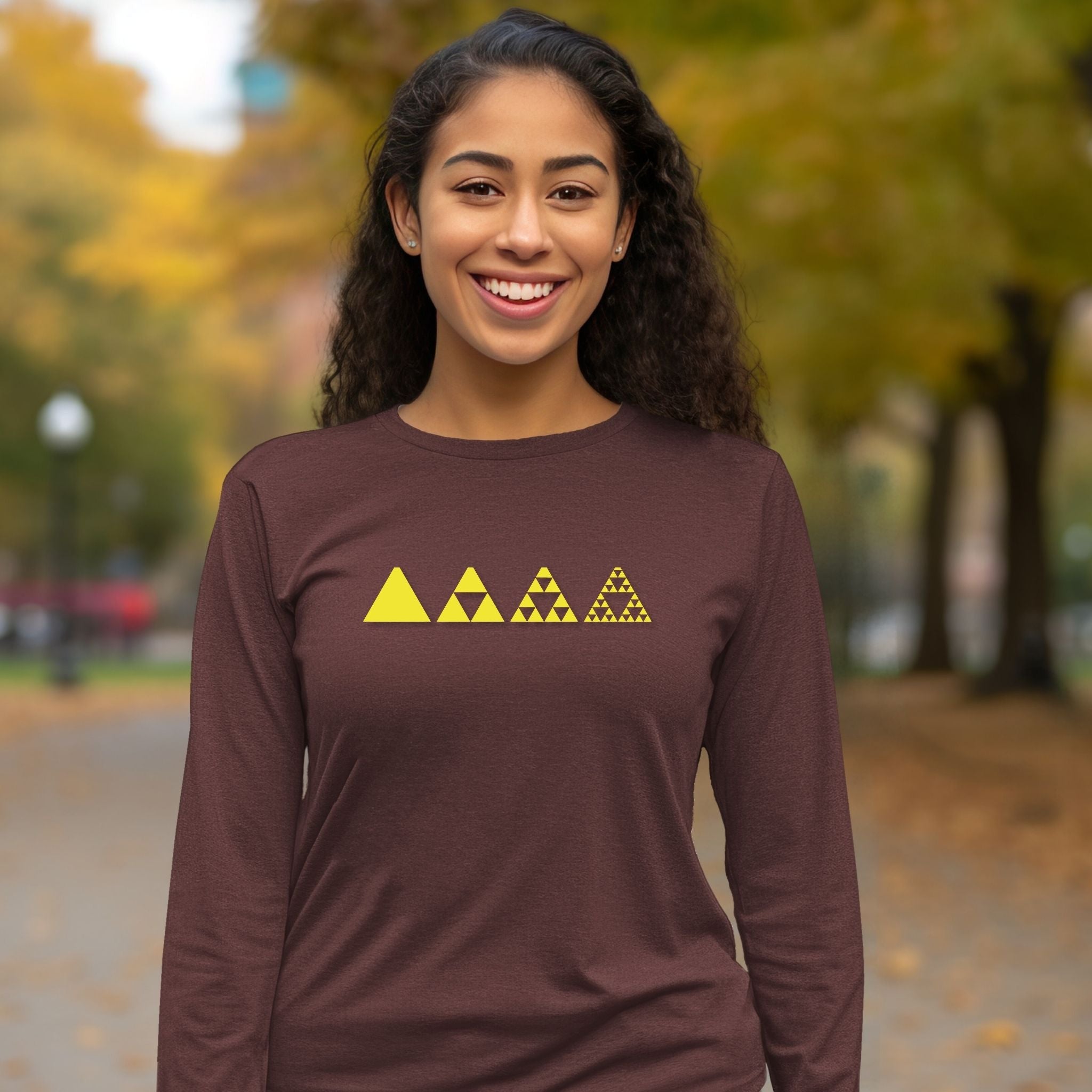 A woman smiling outdoors wearing a dark brown long-sleeve shirt with a yellow Triforce graphic on the chest, set against a blurred autumn park background.