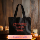 Black tote bag with 'Strange things are happening in this bag' text, pumpkins, and candles in the background.