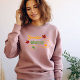 A woman wearing a dusty pink crewneck sweatshirt with a colorful 'Sunday Weather' graphic on the chest, posed against a light wall with a plant in the background.