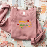 A folded dusty pink long-sleeve sweatshirt with 'Summer Weather' printed in yellow and green with autumn leaves on the chest, styled on a background of fashion magazines.