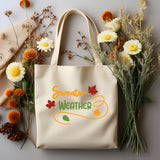 A cream canvas tote bag with 'Sweater WEATHER' printed in orange and green with autumn leaves, styled in a flat lay with dried flowers, pinecones, and a beige scarf on a gray background.