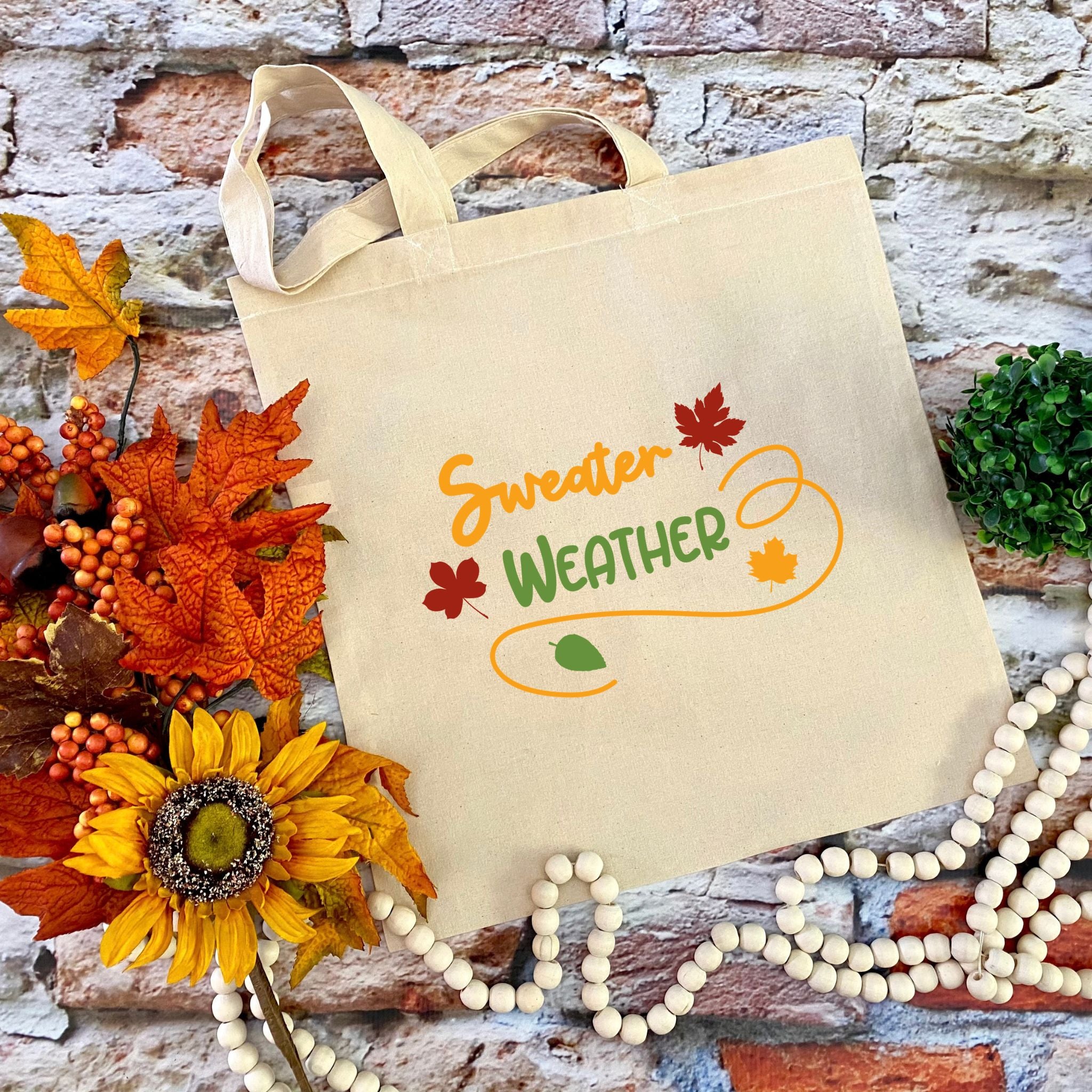 A beige canvas tote bag with 'Sweater WEATHER' printed in orange and green autumn-themed lettering, styled on a brick background with fall leaves, a sunflower, and wooden beads.