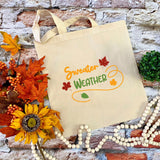 A beige canvas tote bag with 'Sweater WEATHER' printed in orange and green autumn-themed lettering, styled on a brick background with fall leaves, a sunflower, and wooden beads.