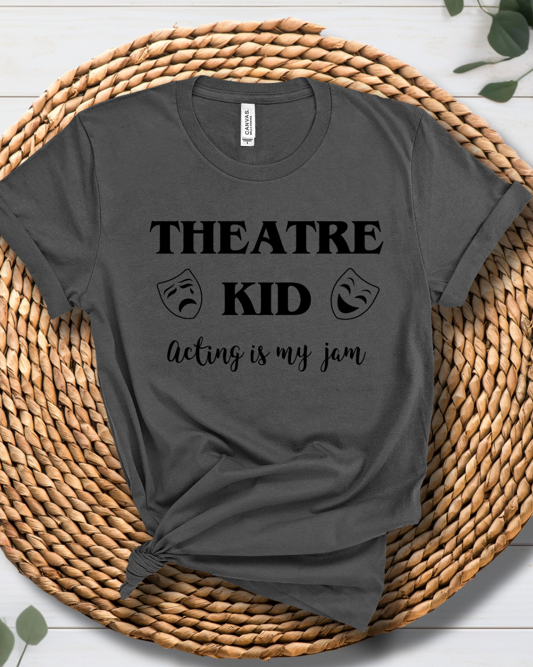 A dark gray graphic t-shirt with black text reading 'THEATRE KID acting is my jam' and theatrical mask icons, styled on a round woven straw mat.