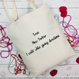 An off-white canvas tote bag with black handwritten text reading 'True New Yorker I walk while giving directions', styled on a white wood background with red rose petals and beaded necklaces.