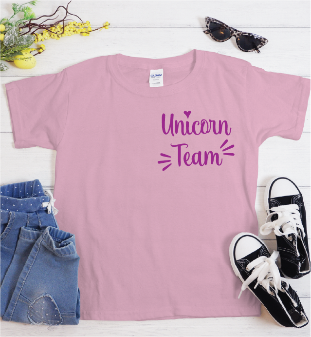 A pink short-sleeve t-shirt with 'Unicorn Team' printed in purple on the front, styled in a flat lay with blue jeans, black sneakers, sunglasses, and yellow flowers on a white wood background.