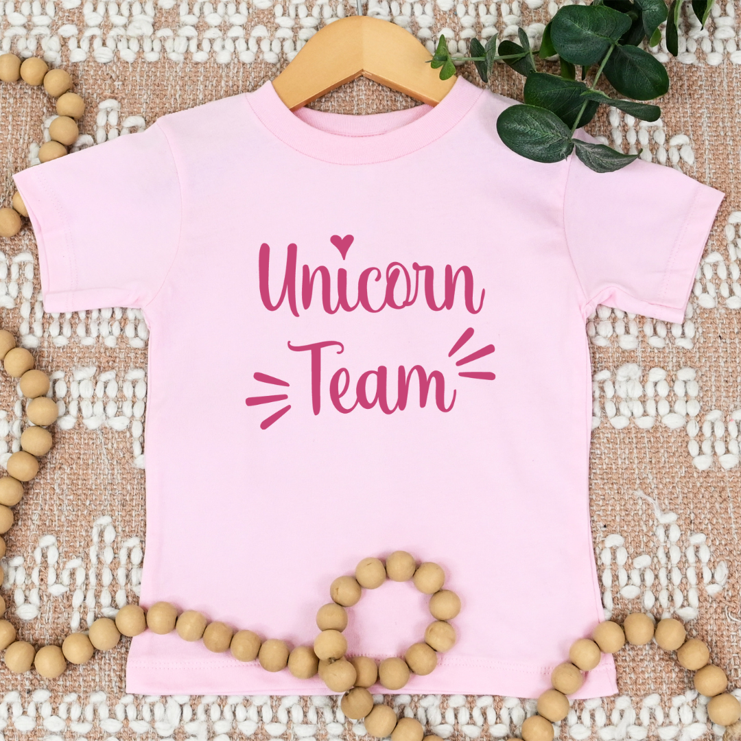 A light pink short-sleeve t-shirt with 'Unicorn Team' printed in dark pink script, displayed on a wooden hanger over a textured beige rug with decorative wooden beads and green leaves.