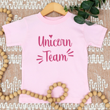 A light pink short-sleeve t-shirt with 'Unicorn Team' printed in dark pink script, displayed on a wooden hanger over a textured beige rug with decorative wooden beads and green leaves.