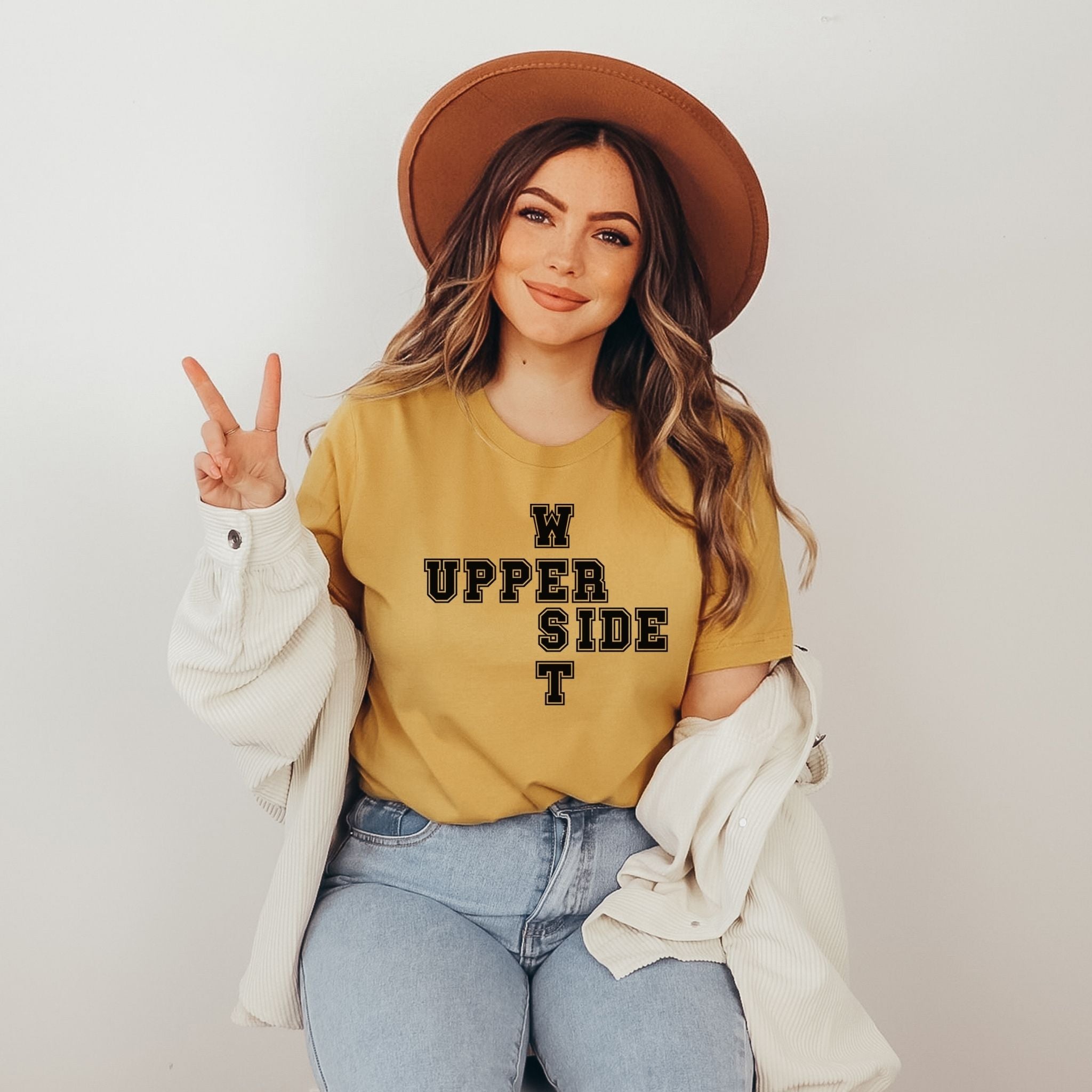 A woman wearing a mustard yellow graphic t-shirt with 'UPPER WEST SIDE' printed in black, styled with a brown wide-brim hat, a white jacket draped off her shoulders, and light blue jeans, posing against a light gray background.