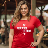 A woman wearing a red short-sleeve t-shirt with white text reading 'UPPER WEST SIDE ' on the front, standing in a blurred outdoor market setting.