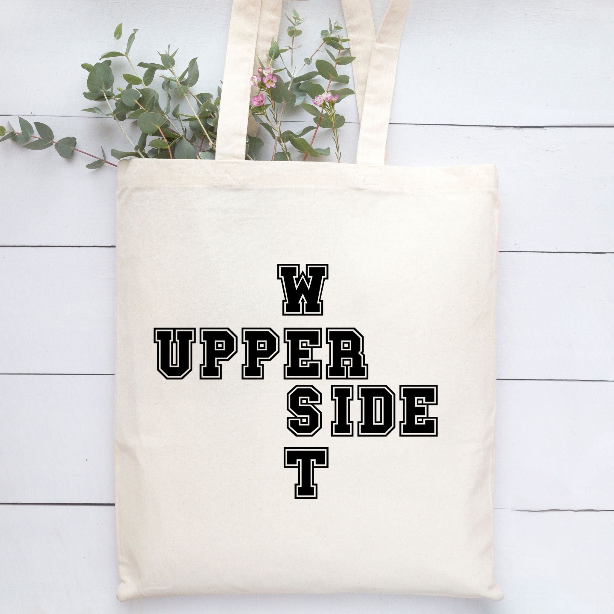 A cream canvas tote bag with black varsity-style block lettering spelling 'Upper West Side' arranged in a cross shape, displayed on a white wood plank background with green eucalyptus stems.