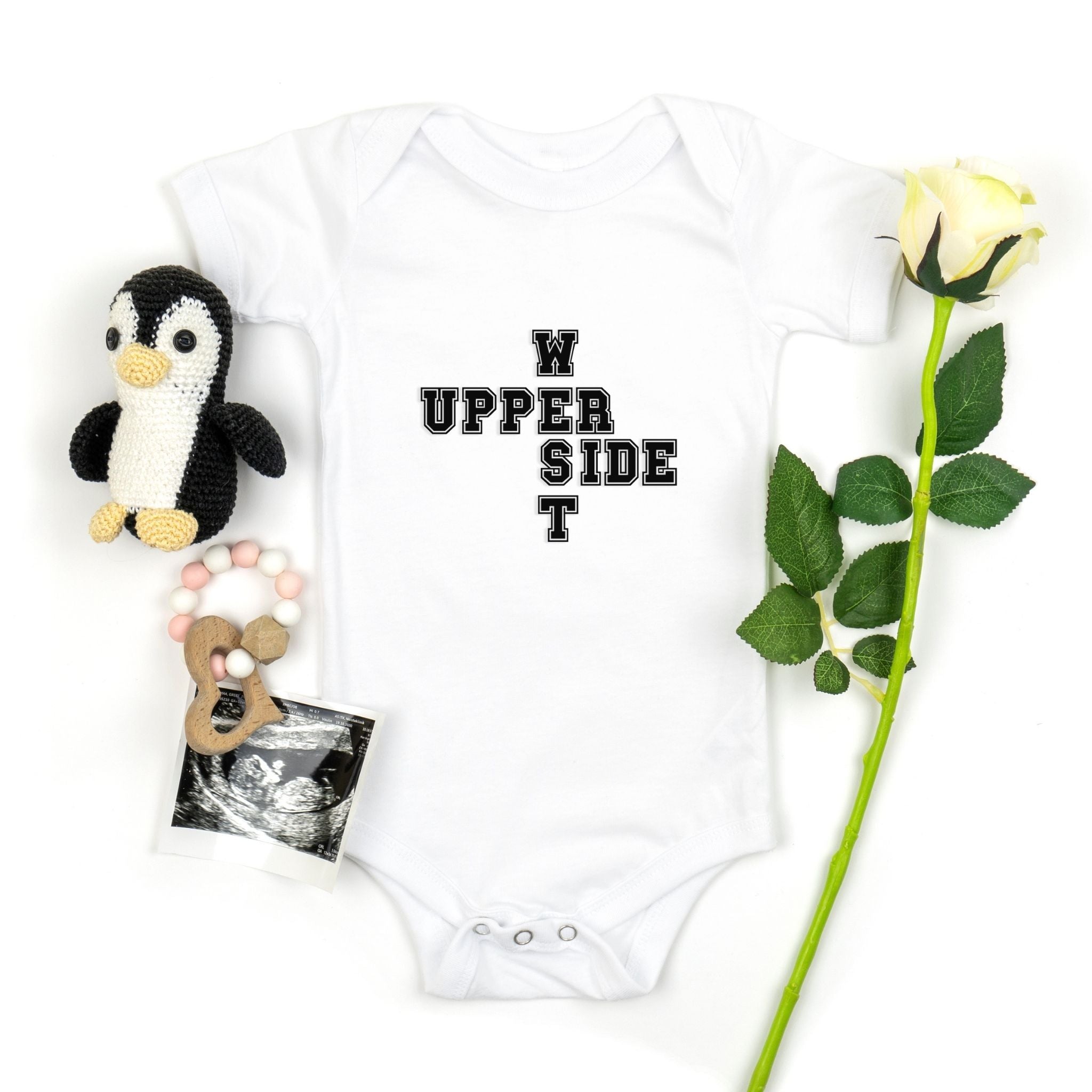 A white baby bodysuit with black text reading 'UPPER WEST SIDE ' laid flat on a white background, styled with a small penguin plush, a baby photo print, and a single white rose.