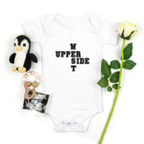 A white baby bodysuit with black text reading 'UPPER WEST SIDE ' laid flat on a white background, styled with a small penguin plush, a baby photo print, and a single white rose.