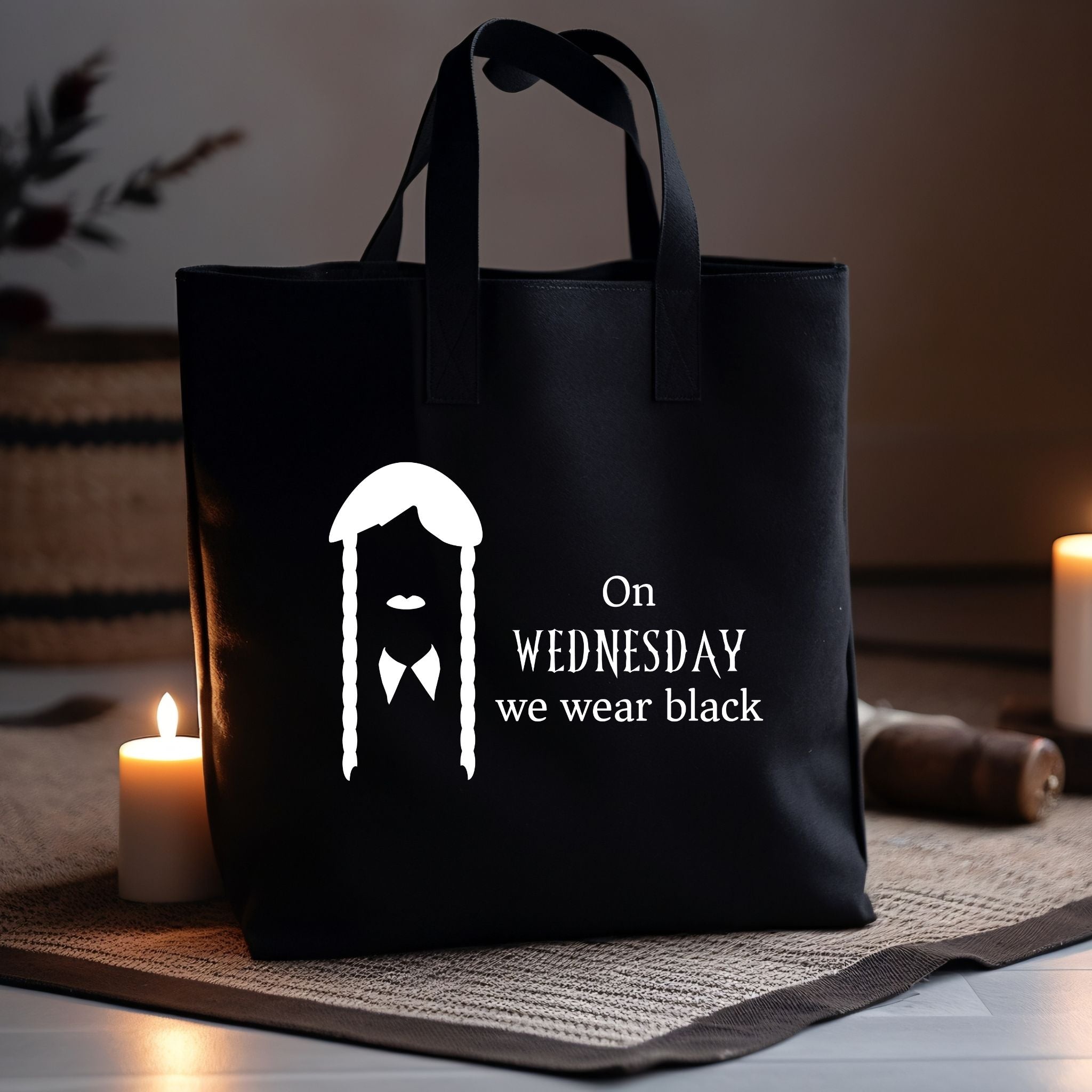 A black tote bag with a white graphic of a man with a mustache and braids, next to the text 'On WEDNESDAY we wear black', displayed on a woven mat with lit candles in a warm, dimly lit setting.