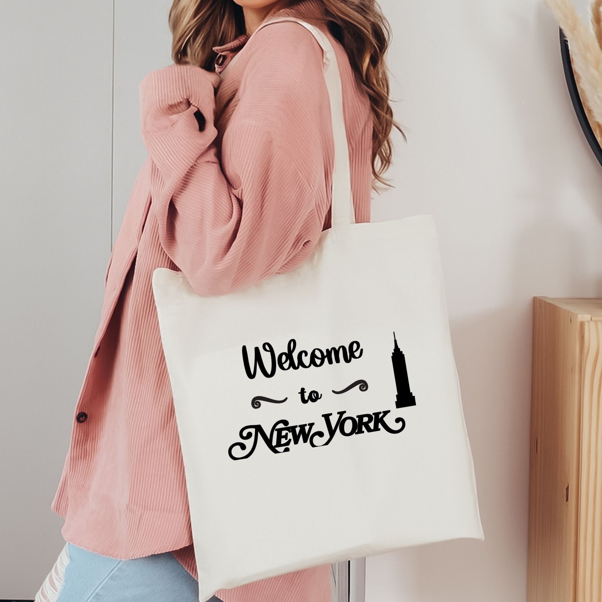 A woman wearing a pink cardigan carries a white canvas tote bag with black text reading 'Welcome to New York' and a small Empire State Building silhouette.