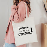 A woman wearing a pink cardigan carries a white canvas tote bag with black text reading 'Welcome to New York' and a small Empire State Building silhouette.