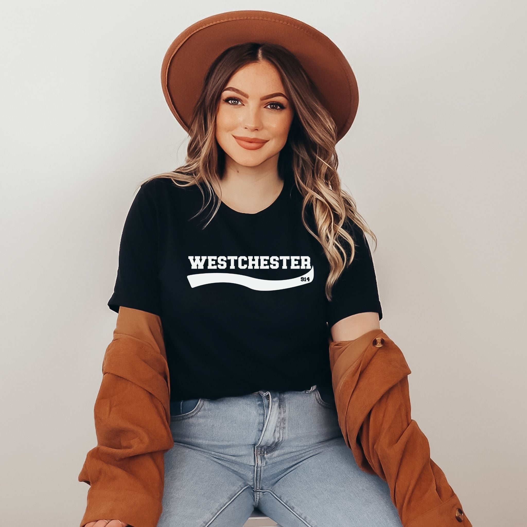 A woman wearing a black t-shirt with 'WESTCHESTER' printed in white across the chest, styled with a brown wide-brim hat, a brown jacket draped off her shoulders, and light blue jeans against a plain light gray background.