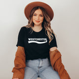 A woman wearing a black t-shirt with 'WESTCHESTER' printed in white across the chest, styled with a brown wide-brim hat, a brown jacket draped off her shoulders, and light blue jeans against a plain light gray background.