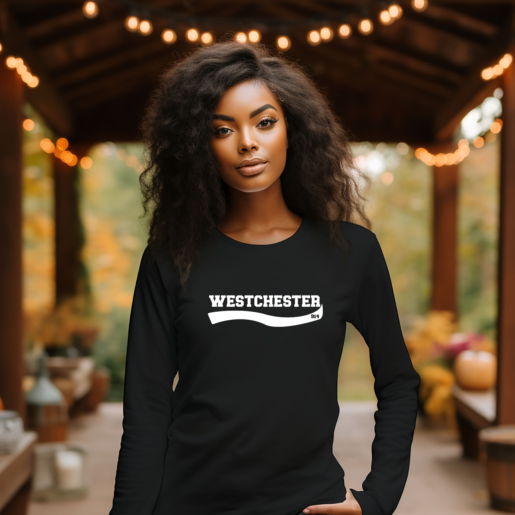 A woman with curly black hair stands under a wooden covered patio wearing a black long-sleeve shirt with a white 'WESTCHESTER' graphic across the chest. The background features warm string lights, autumn foliage, and pumpkins.