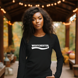 A woman with curly black hair stands under a wooden covered patio wearing a black long-sleeve shirt with a white 'WESTCHESTER' graphic across the chest. The background features warm string lights, autumn foliage, and pumpkins.