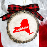 A circular wood slice ornament with a red silhouette of New York and the word 'Westchester' on it, hanging from a red and black plaid ribbon bow.