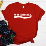 A red short-sleeve T-shirt laid flat on a white wood background, with 'WESTCHESTER 914' printed in white across the chest. The shirt is styled with a floral fabric prop at the lower left.