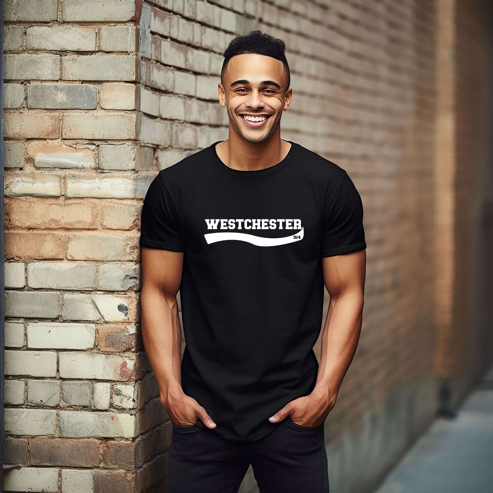 A smiling man wearing a black t-shirt with 'WESTCHESTER 914' printed in white across the chest, standing against a brick wall.