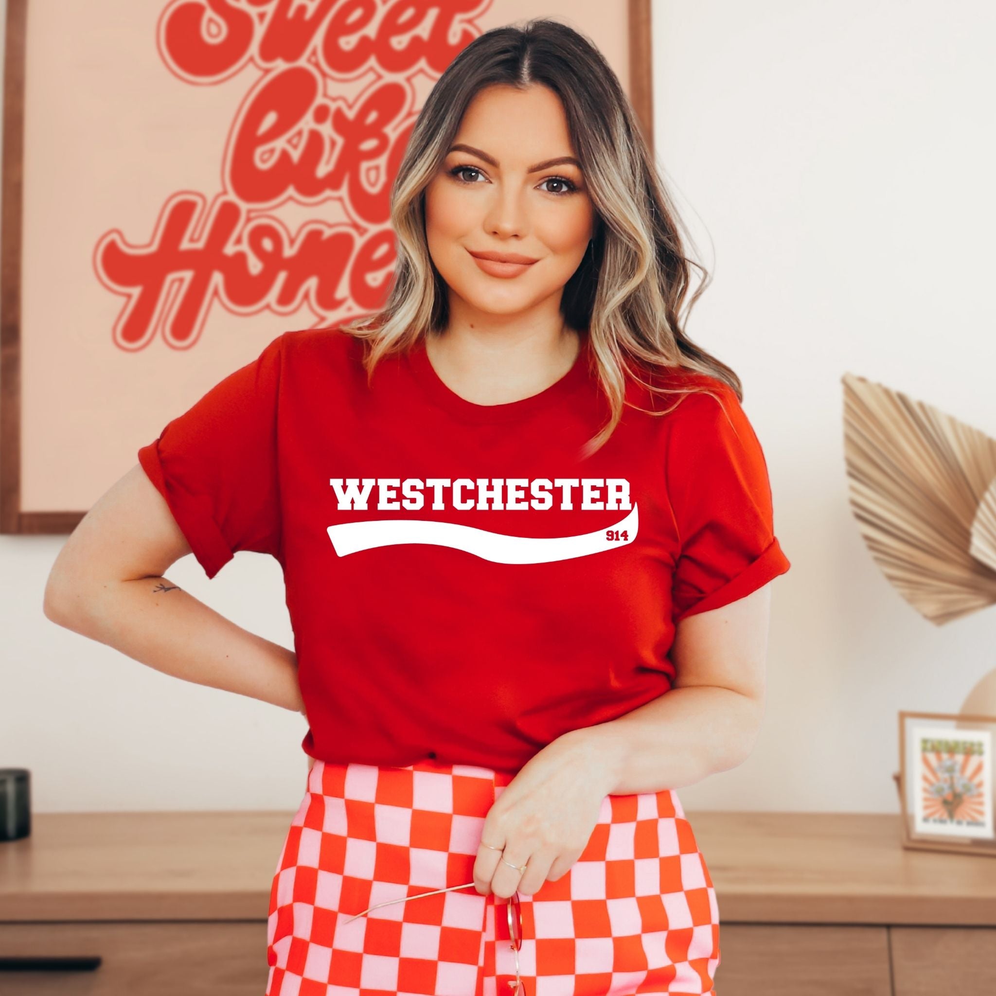 A woman wearing a red short-sleeve t-shirt with 'WESTCHESTER' printed in white across the chest, paired with red and white checkered shorts, standing in a decorated indoor setting.